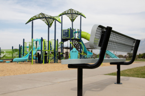 A metal bench in the foreground with the Mountain Ridge Park playground, including slides and climbing structures, in the background.